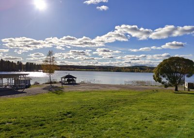 Järven ranta, sinisellä taivaalla valkoisia pilviä, nurmikko, aurinko paistaa.